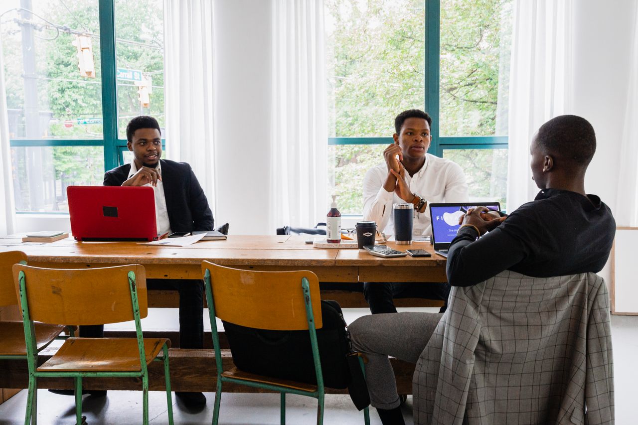 Three men engaged in a work meeting around a wooden table in a bright office.
